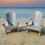Two gray Adirondack chairs with a small  Tete table on a sandy beach.