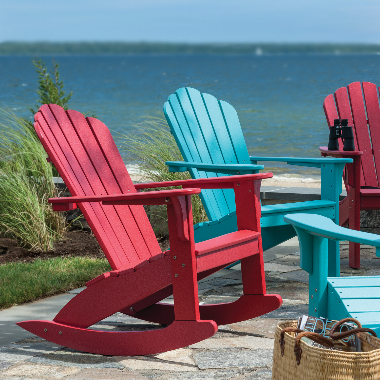 Two red and one blue Adirondack chair by a body of water.