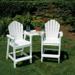 Two white Adirondack classic bar chairs with a table placed on grass against a backdrop of plants and a stone wall.