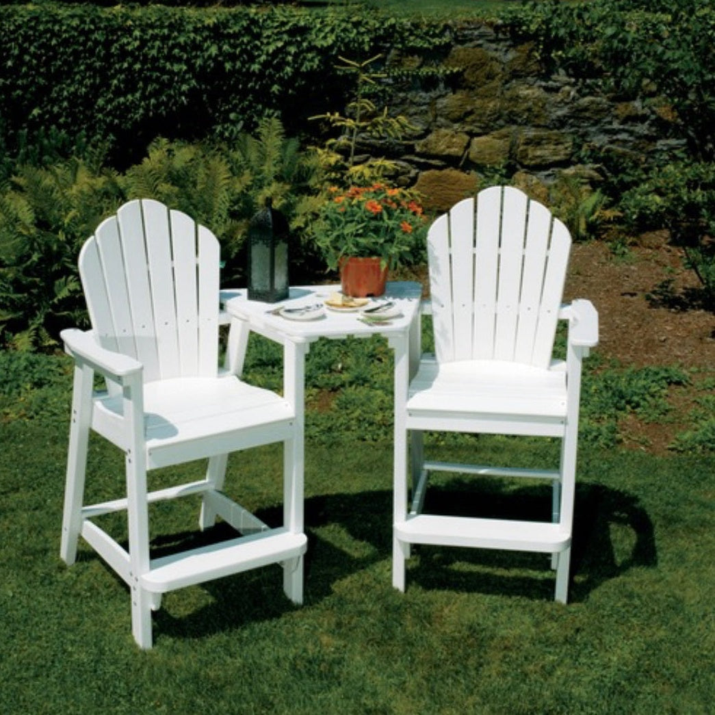 Two white Adirondack classic bar chairs with a table placed on grass against a backdrop of plants and a stone wall.
