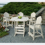 A beige square Portsmouth bar table with four classic Adirondack bar chairs set on a gravel surface, with plants and a bottle of wine visible in the background.