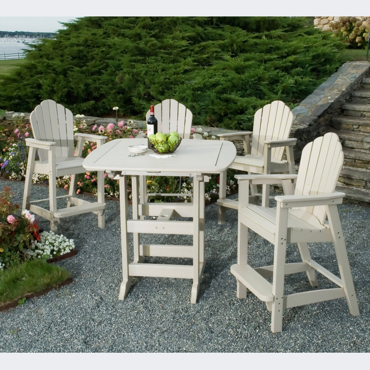 A beige square Portsmouth bar table with four classic Adirondack bar chairs set on a gravel surface, with plants and a bottle of wine visible in the background.