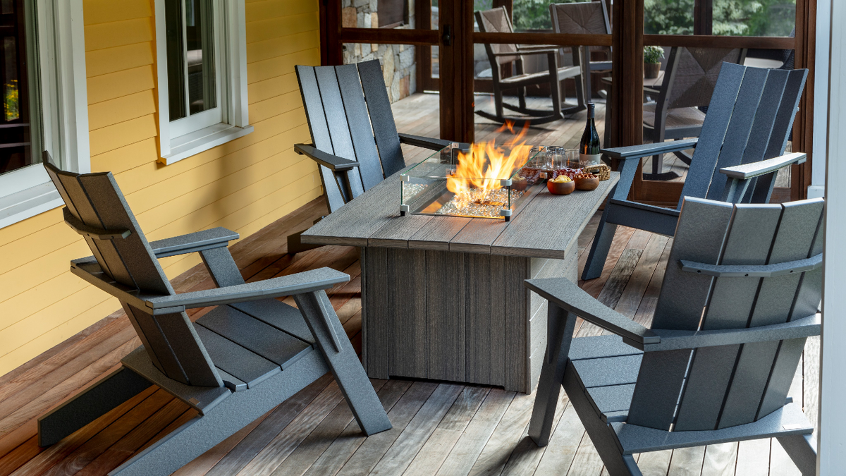 A gray outdoor patio fire pit table surrounded by four matching gray Adirondack-style chairs on a wooden porch, with a visible flame burning in the center of the table.